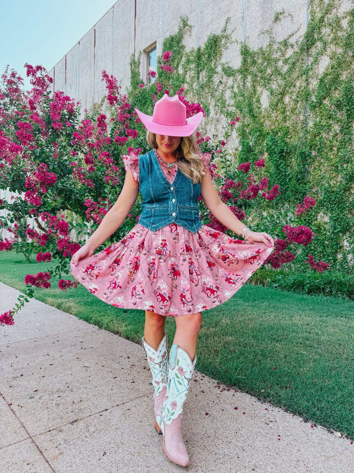 Woman in pink floral dress, denim vest, and pink cowboy boots standing outdoors.