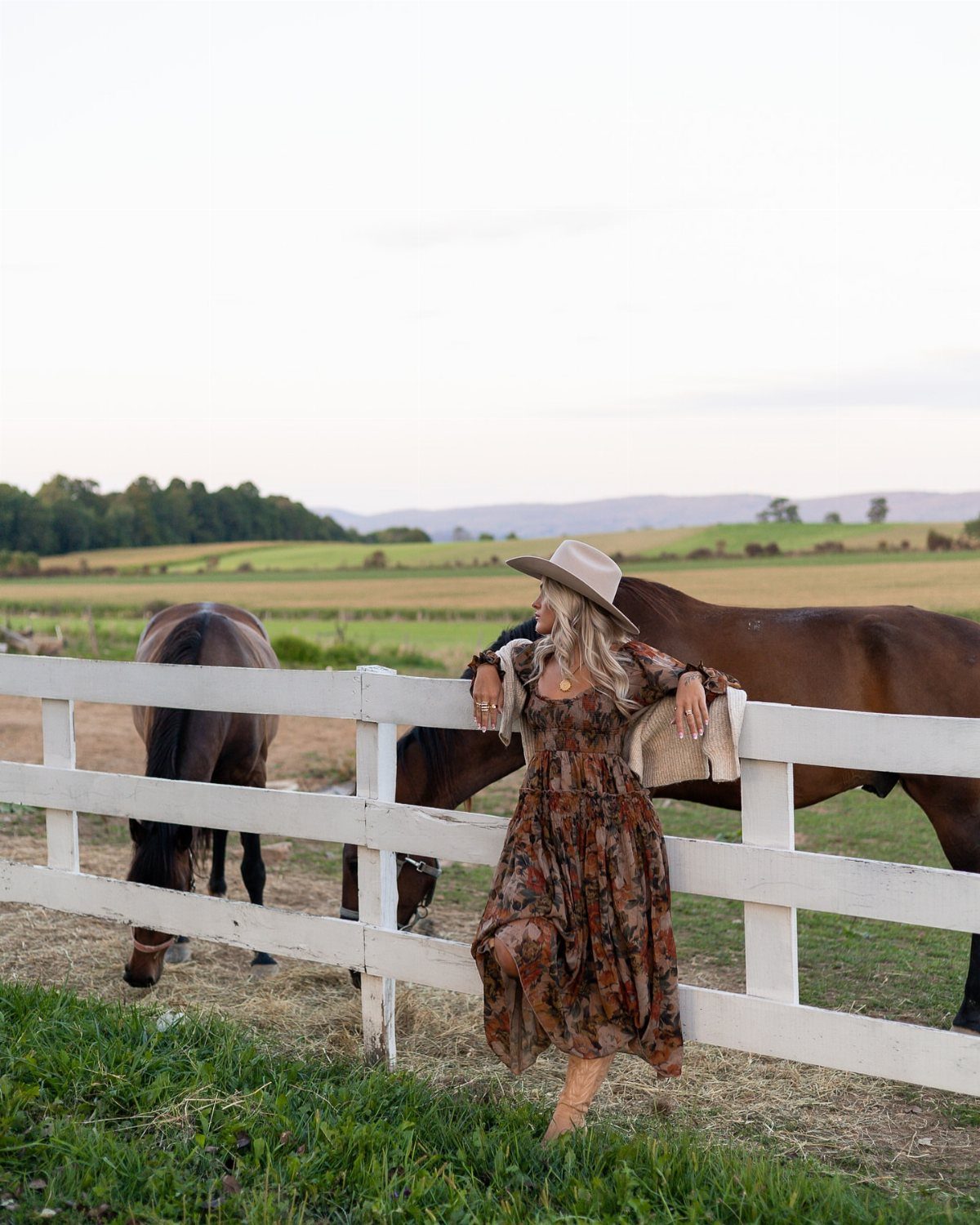 Woman in a brown floral maxi dress leaning against a white fence in a grassy field.