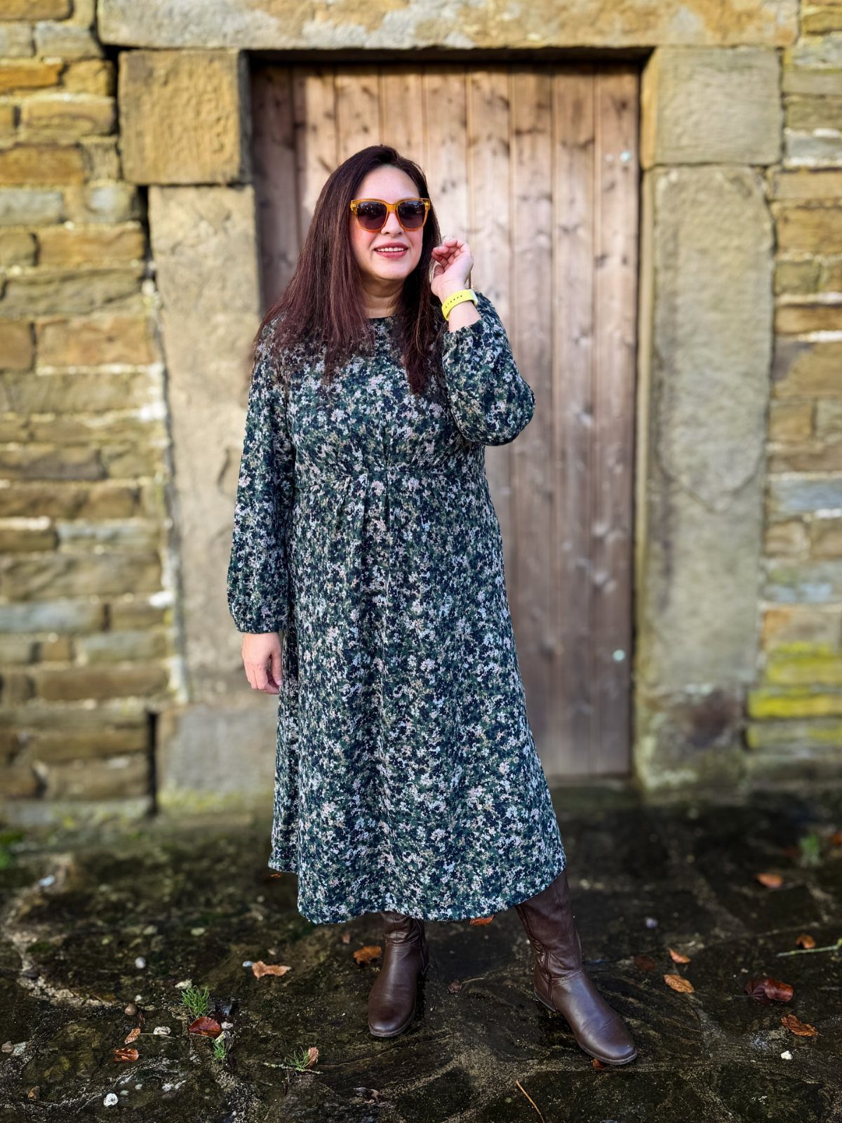 A woman wearing a floral midi dress and brown leather boots standing before a wooden door.