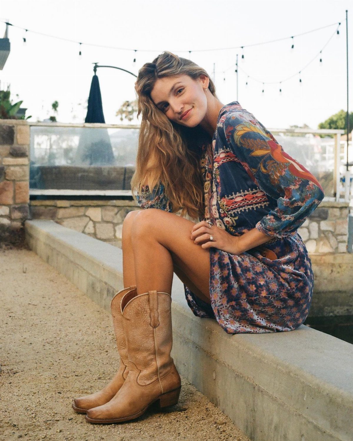 A woman sitting on a ledge wearing a patterned flowy dress and tan cowboy boots.