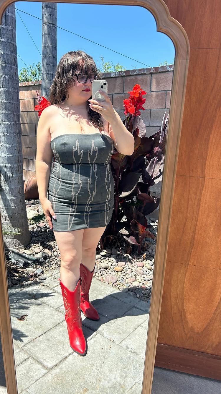 A woman in a short grey strapless dress and red cowboy boots taking a mirror selfie.
