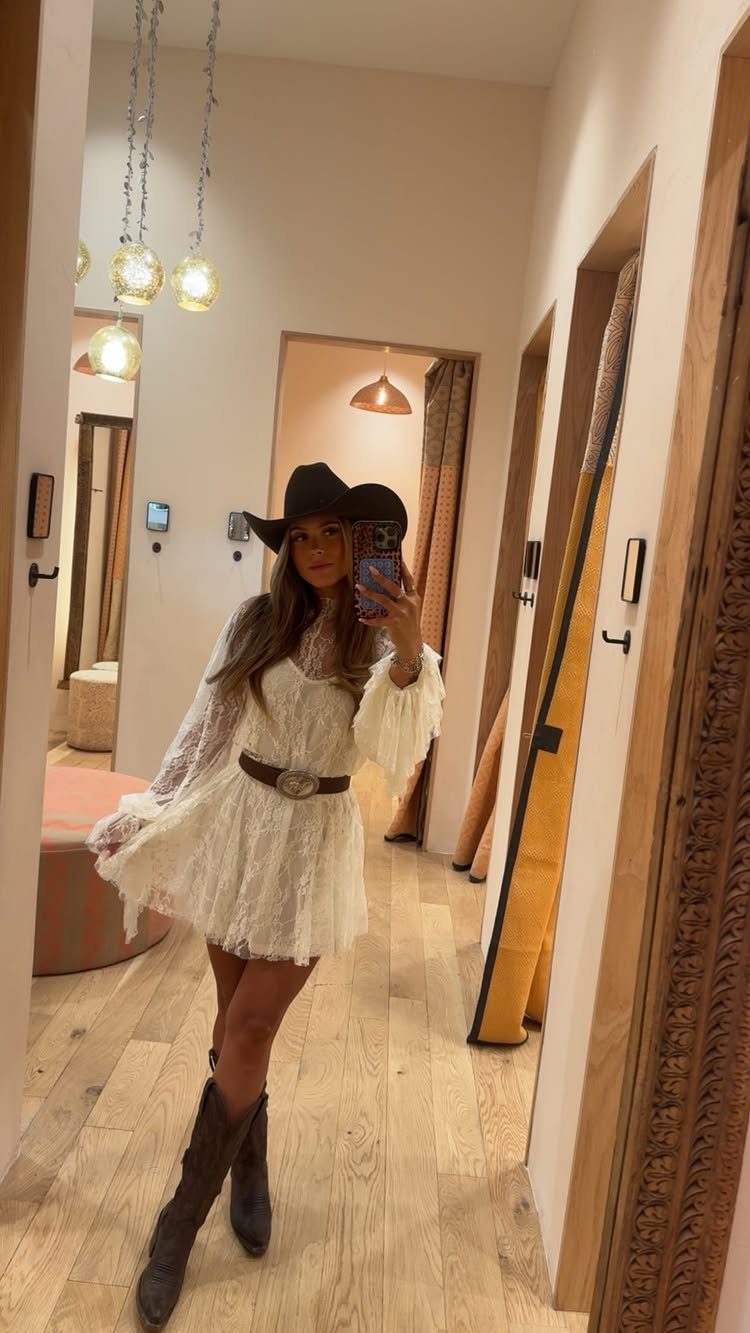 Woman in a white lace dress, cowboy hat, and brown boots posing for a mirror selfie.
