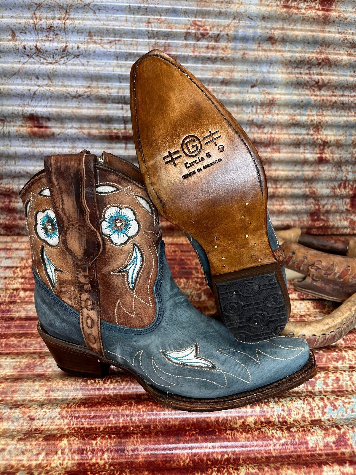 Blue and brown leather western ankle boots with white floral embroidery on a textured background.