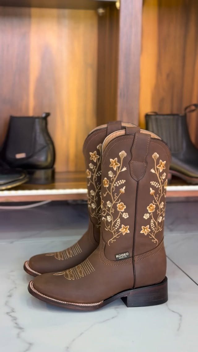 Brown leather cowboy boots with yellow floral embroidery sitting on a white marble floor.