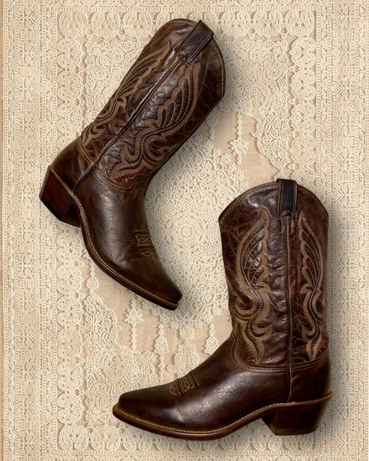 Two brown leather cowboy boots with decorative stitching resting on a patterned beige rug.