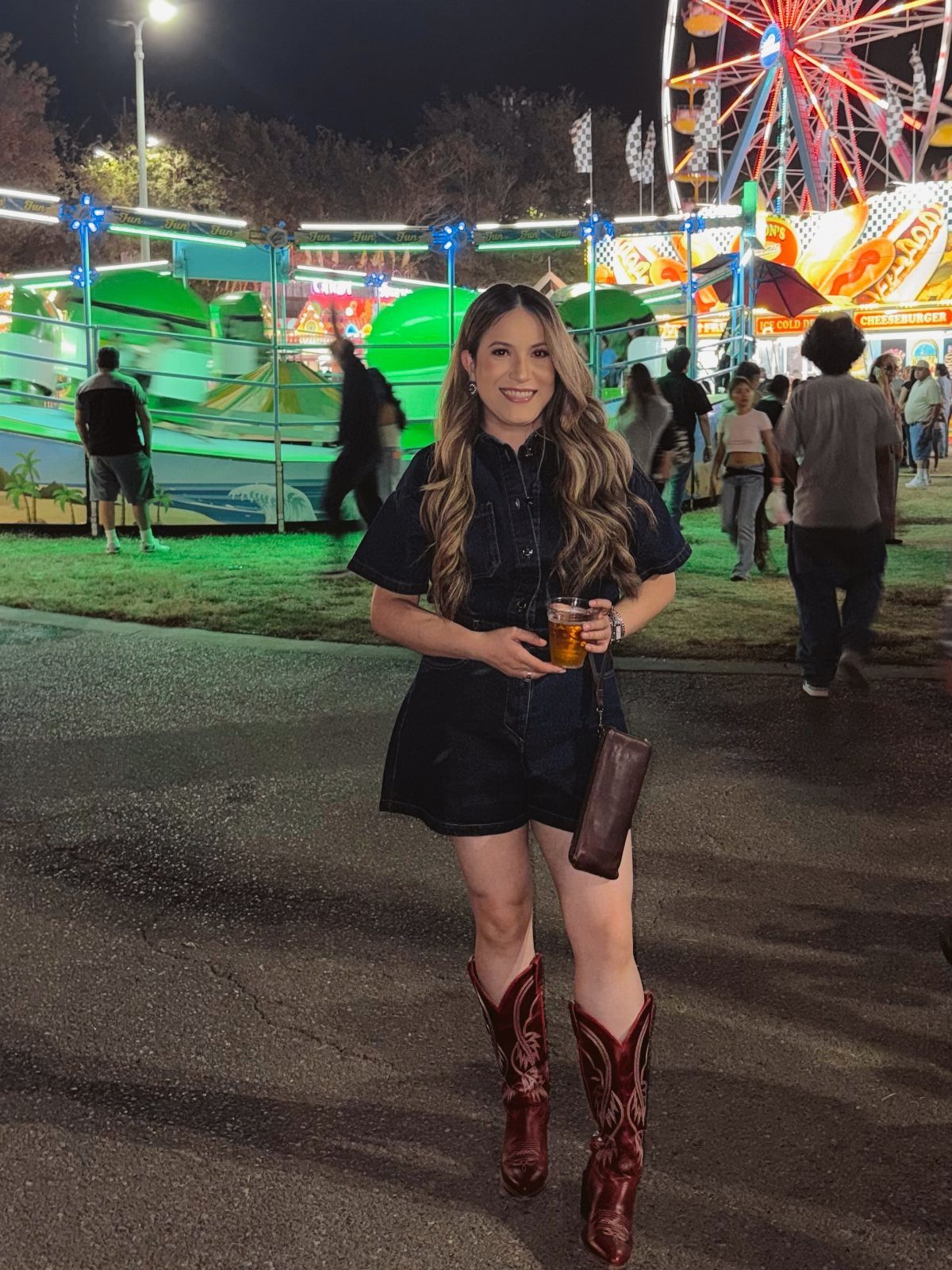 A woman wearing a dark denim romper and cowboy boots at a night carnival.