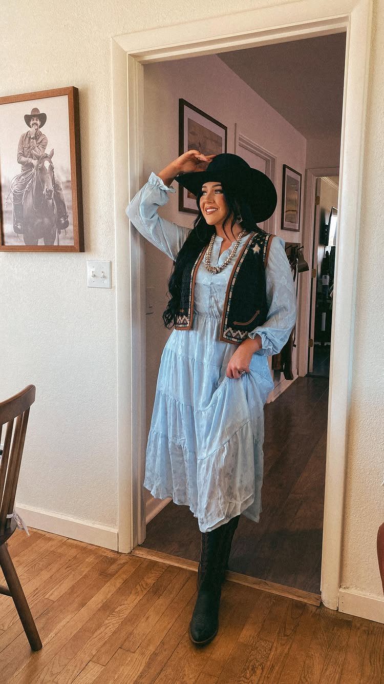 Woman in a light blue midi dress, black vest, and cowboy boots posing in a doorway.