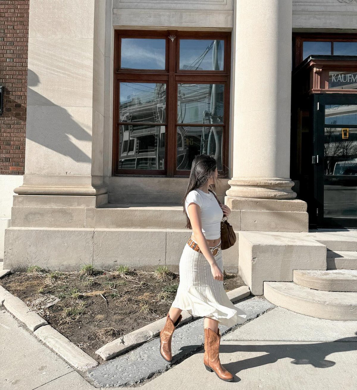 Woman walking in a white t-shirt, flowy white skirt, and brown cowboy boots.
