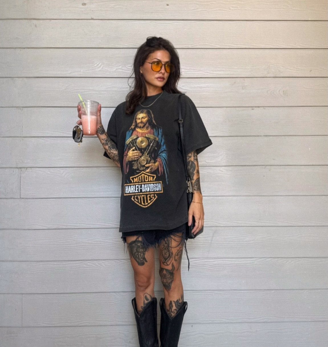 Woman in a black Harley-Davidson graphic tee and black cowboy boots standing against a wooden wall.