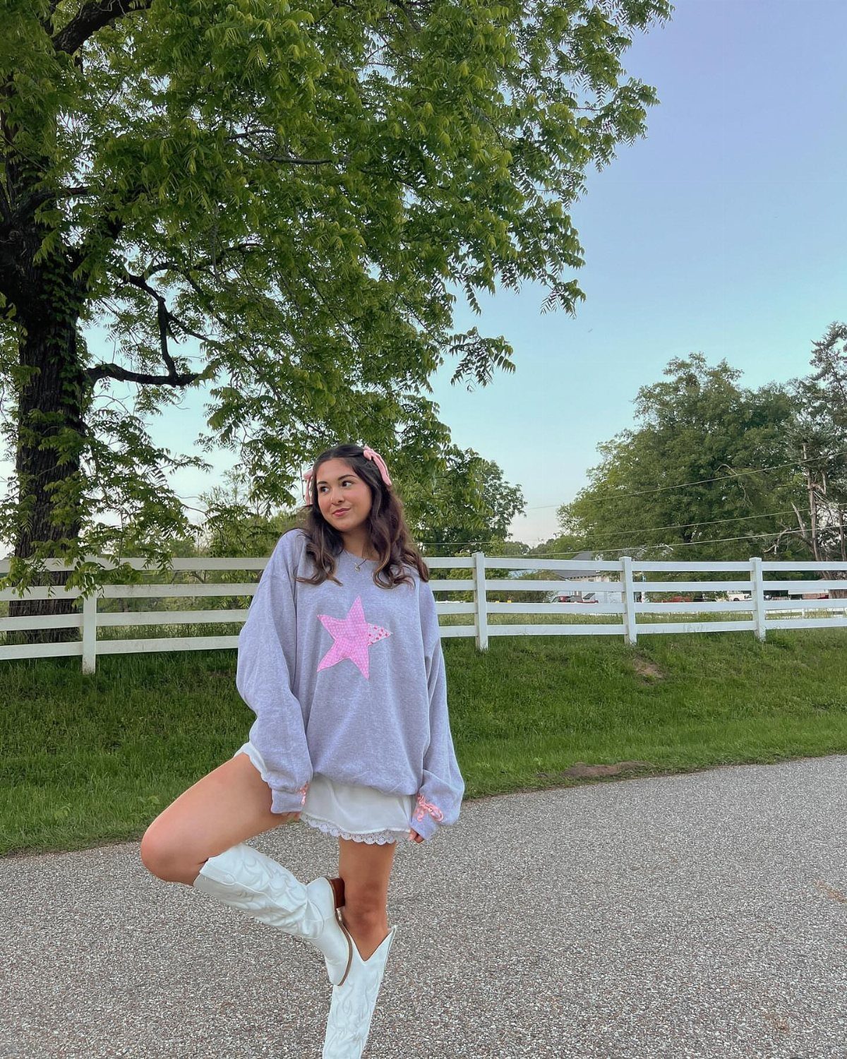 Woman in a purple star sweater, white skirt, and white cowboy boots standing outdoors.