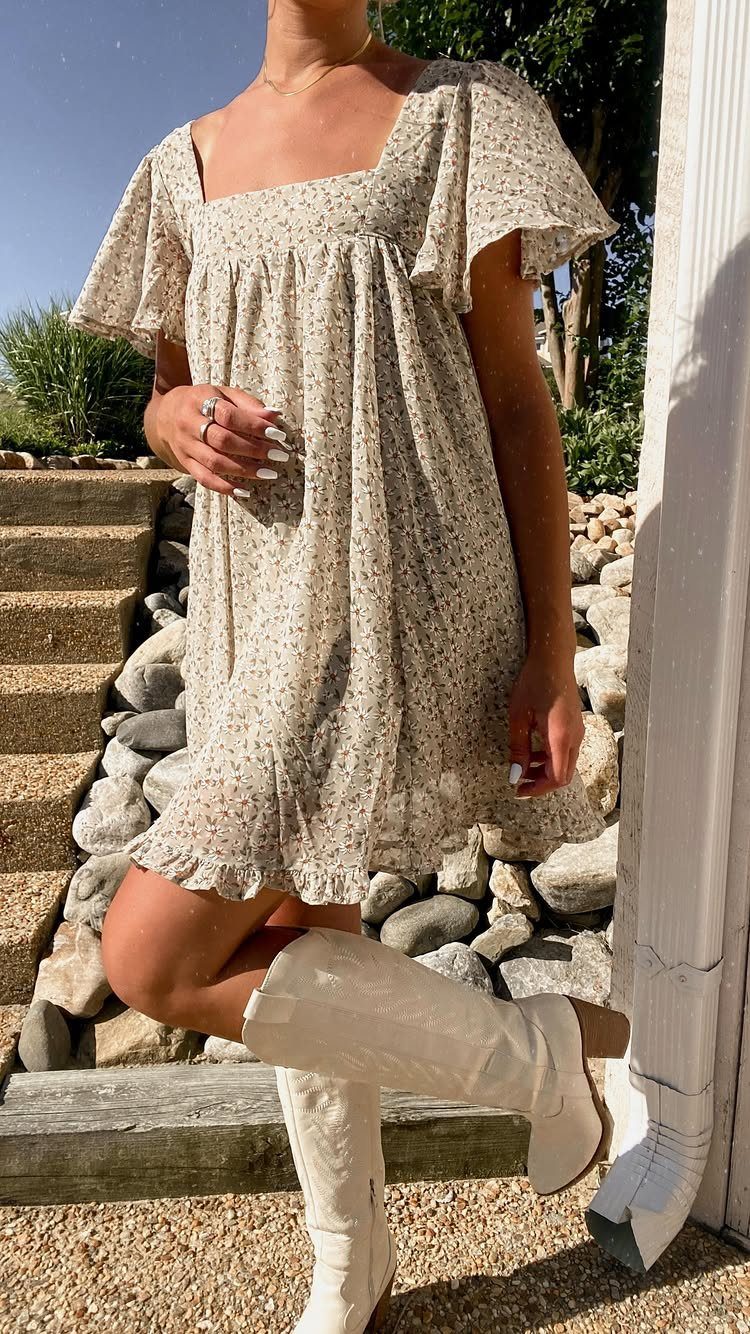 A person wearing a floral mini dress paired with tall white cowboy boots standing outdoors.