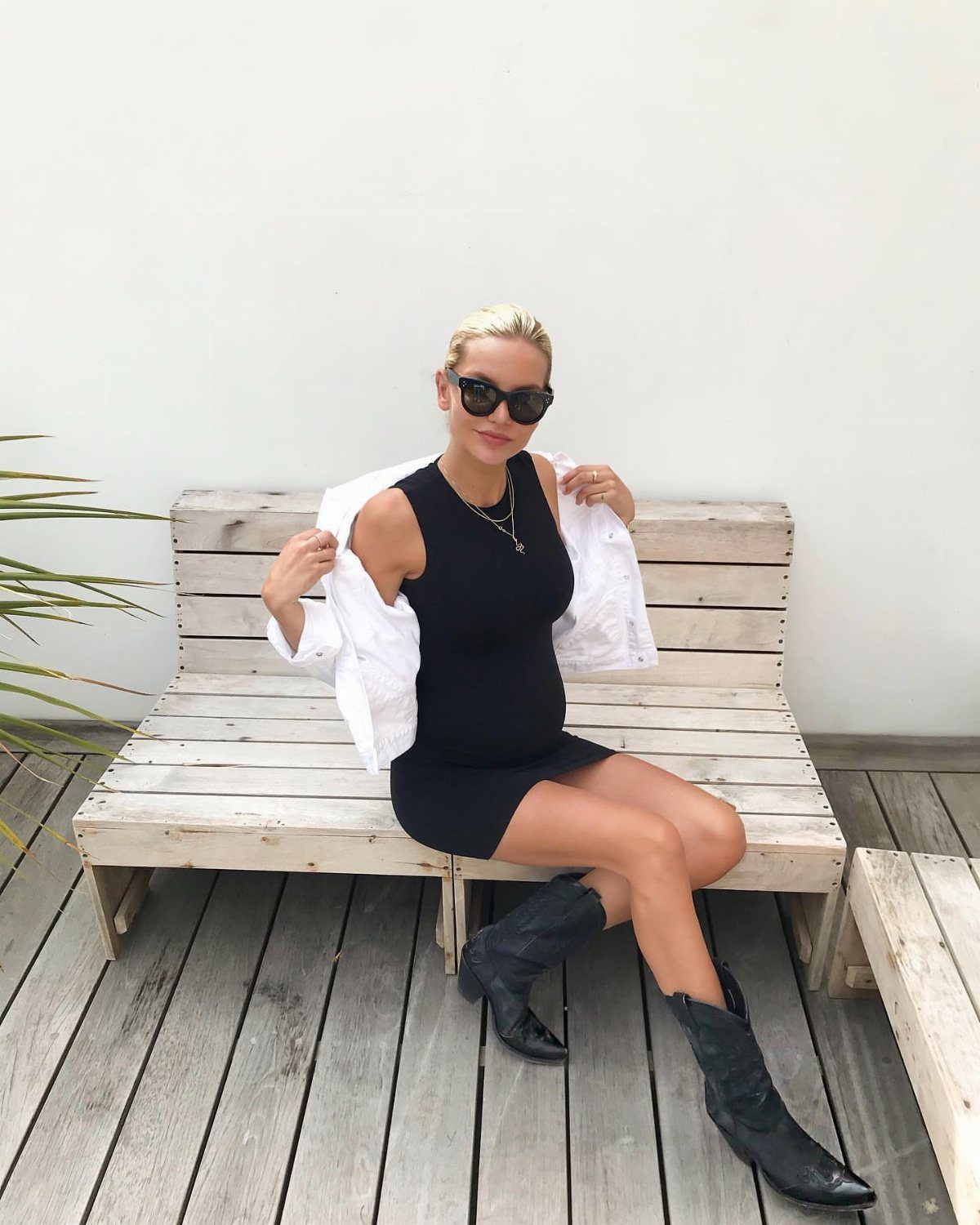 A woman in a black dress and black cowboy boots sitting on a wooden bench.