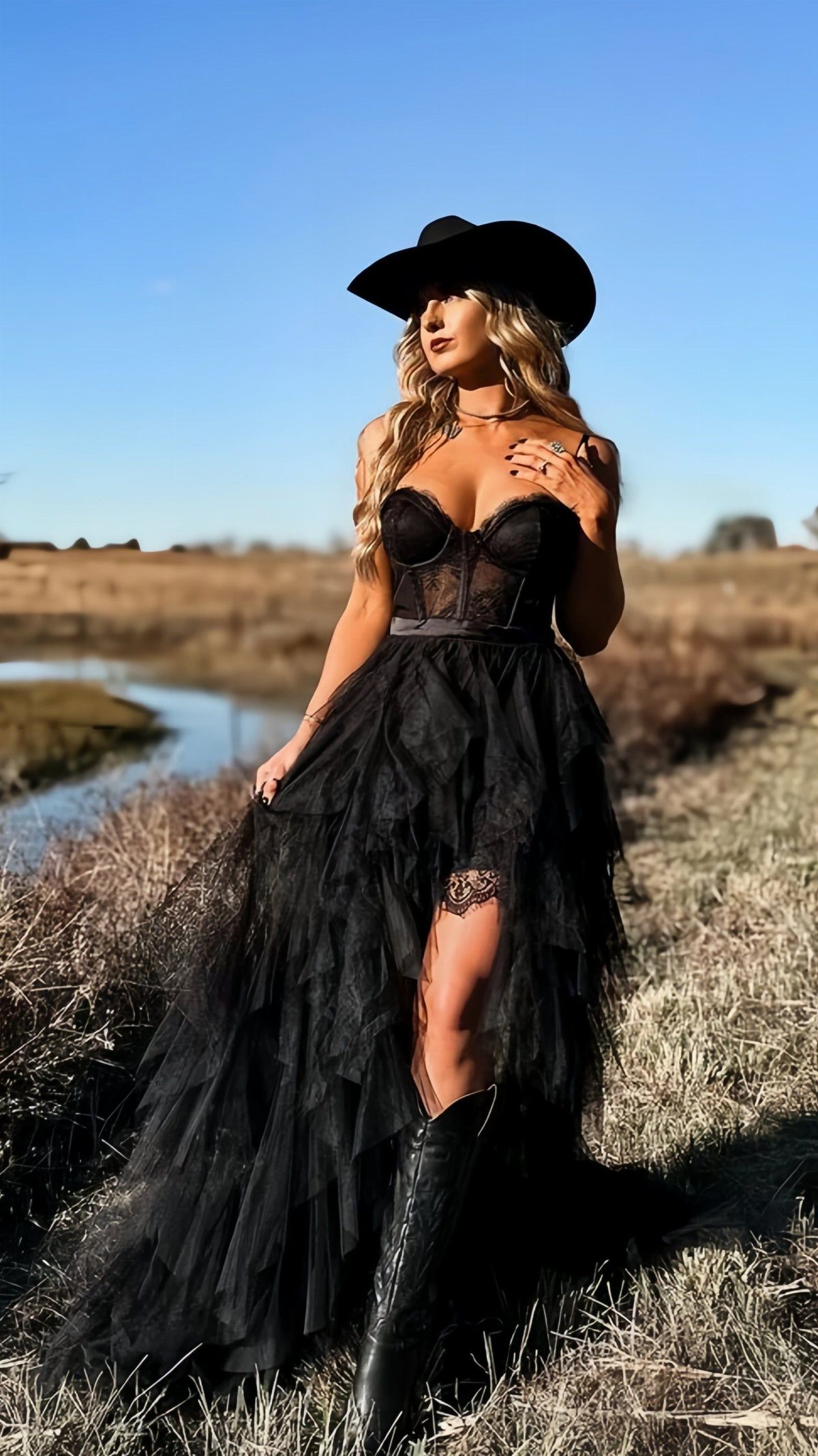 A woman in a black tulle dress and cowboy hat posing in a grassy field.