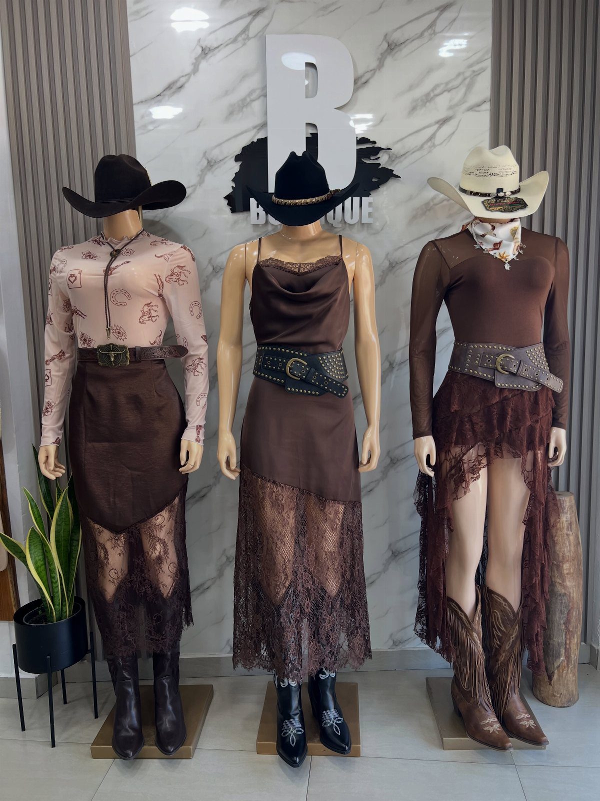 Three mannequins wearing various western-themed brown dresses and cowboy boots against a marble wall.