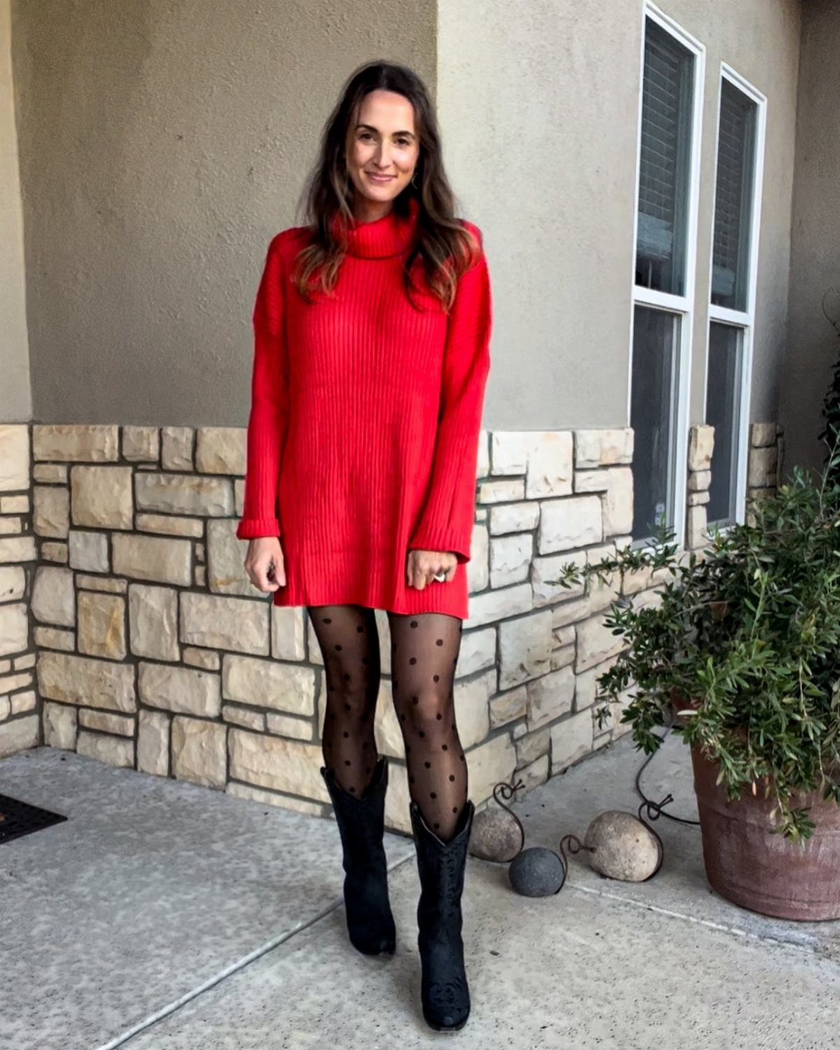 Woman wearing a red knitted sweater dress with black patterned tights and black cowboy boots.