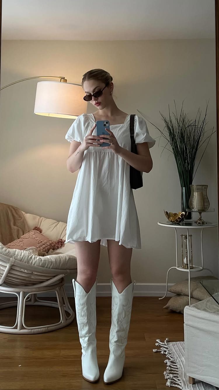 Woman in a white puff-sleeve dress and white cowboy boots taking a mirror selfie.