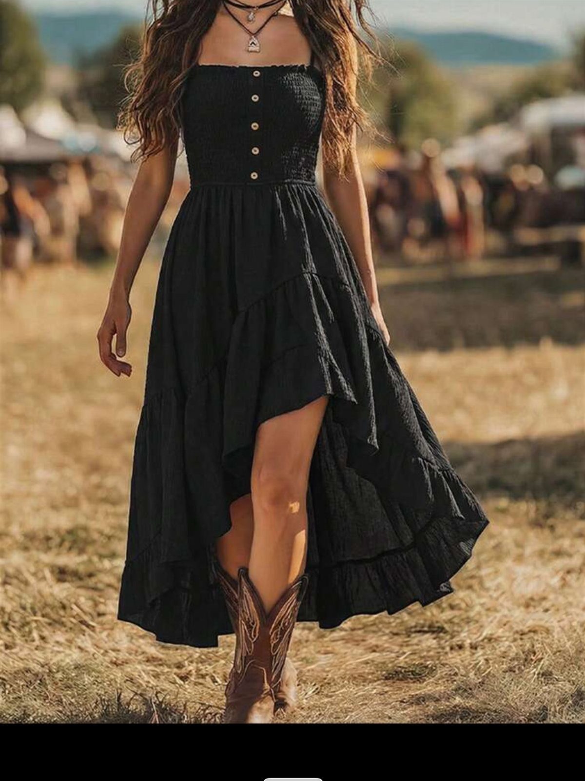 Woman in a black high-low midi dress and brown western boots walking through a field.