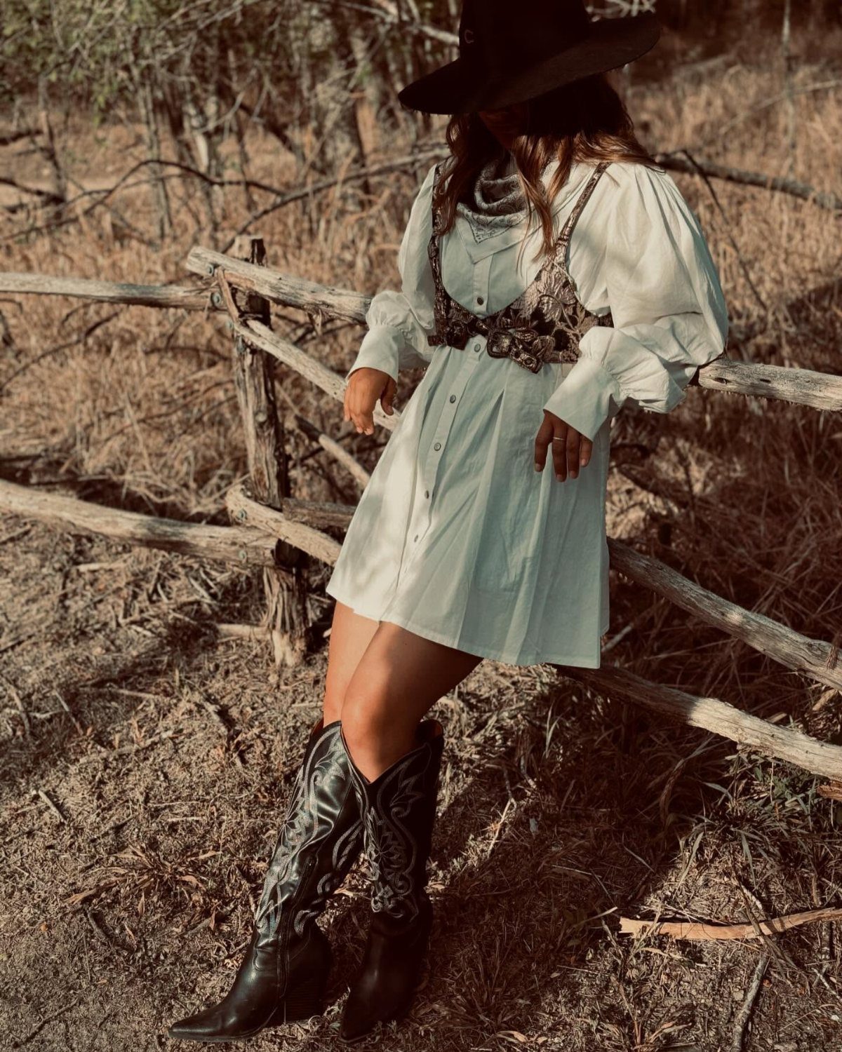 Woman in a white puff sleeve dress and black western boots sitting on a wooden fence.