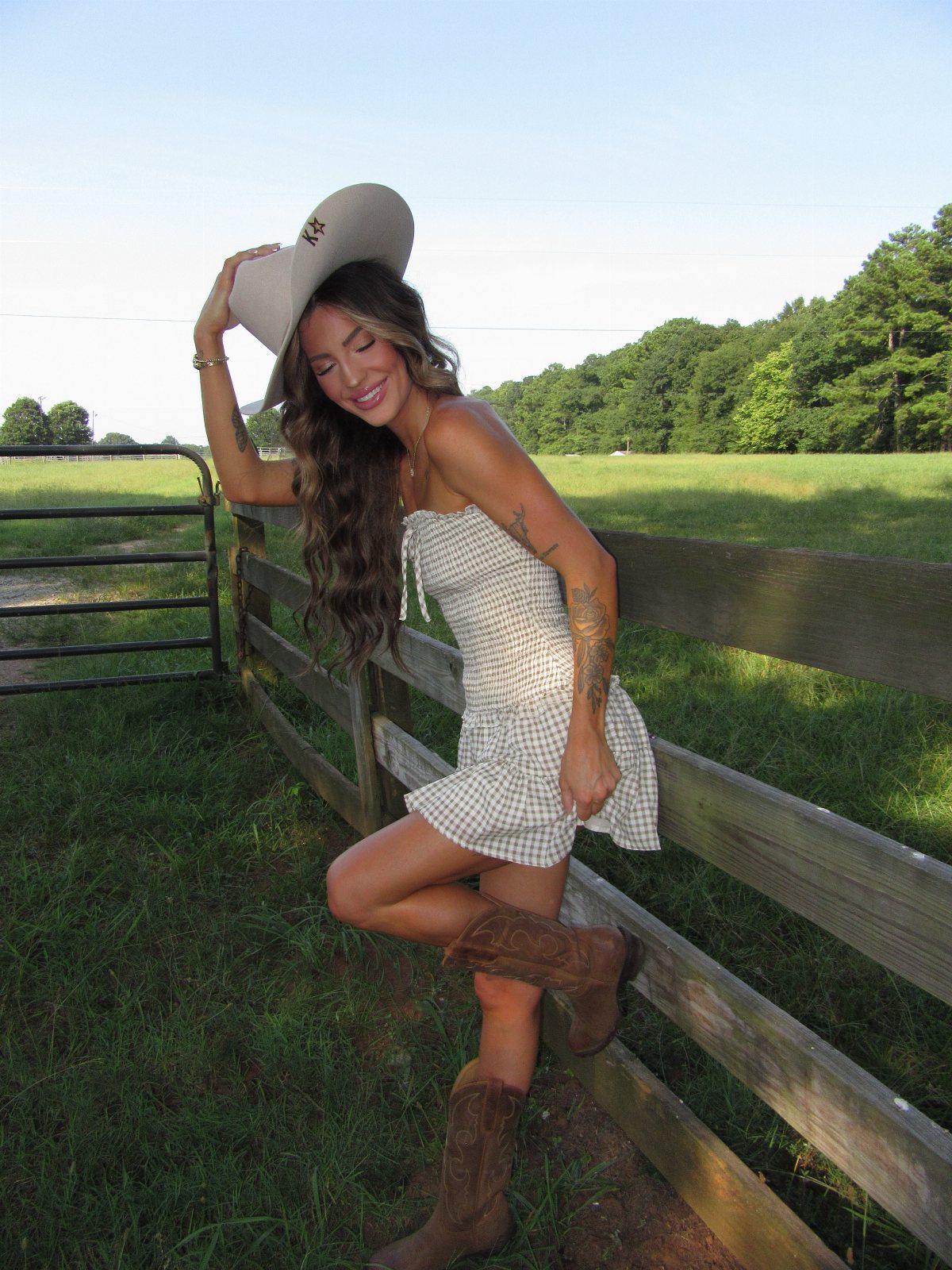 A woman in a white smocked dress and brown western boots posing by a wooden fence.