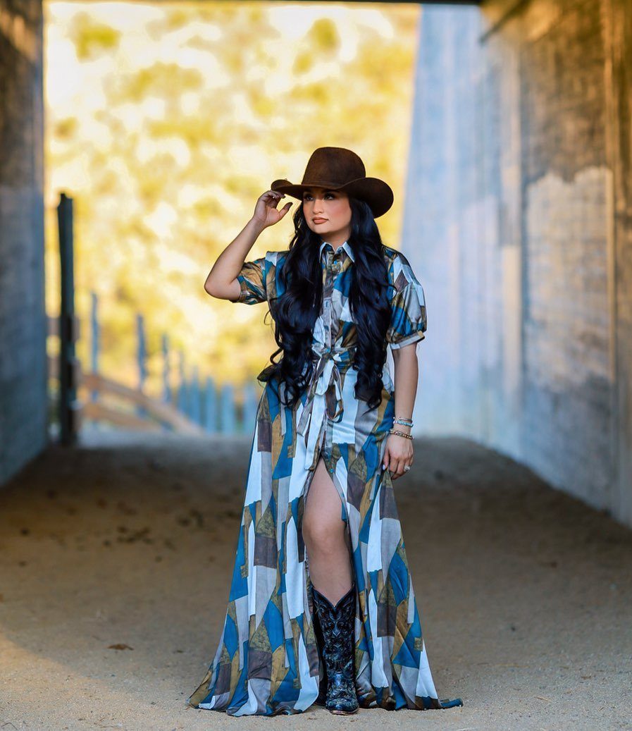 A woman in a patterned maxi dress and cowboy hat posing in a sunlit walkway.