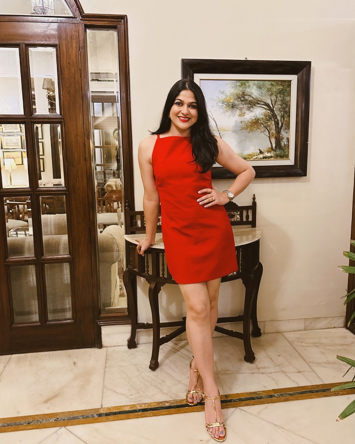 A woman in a red dress and gold strappy heels posing indoors near a wooden cabinet.