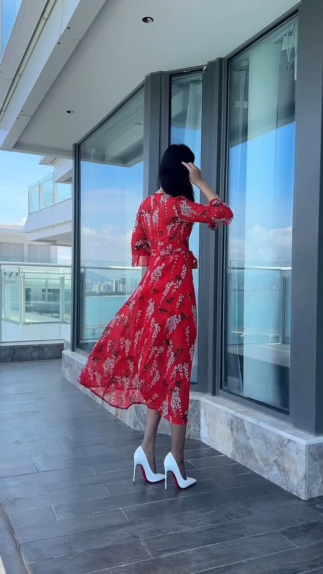 Woman in a red floral dress and white high heels standing on a balcony by the ocean.