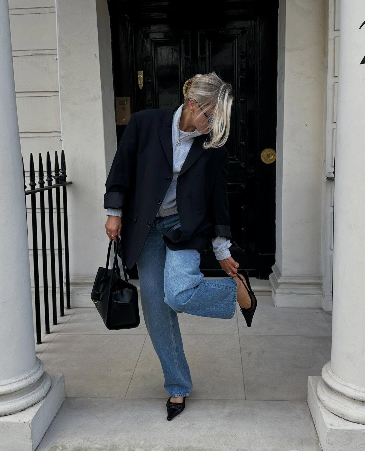 Woman wearing blue jeans, black blazer, and black high heels standing by a doorway.