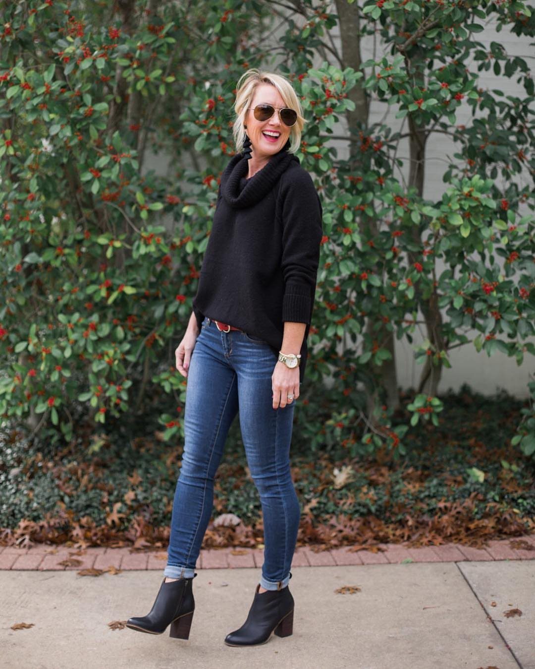 A woman wearing a black sweater, cuffed skinny jeans, and black heeled ankle boots standing outdoors.