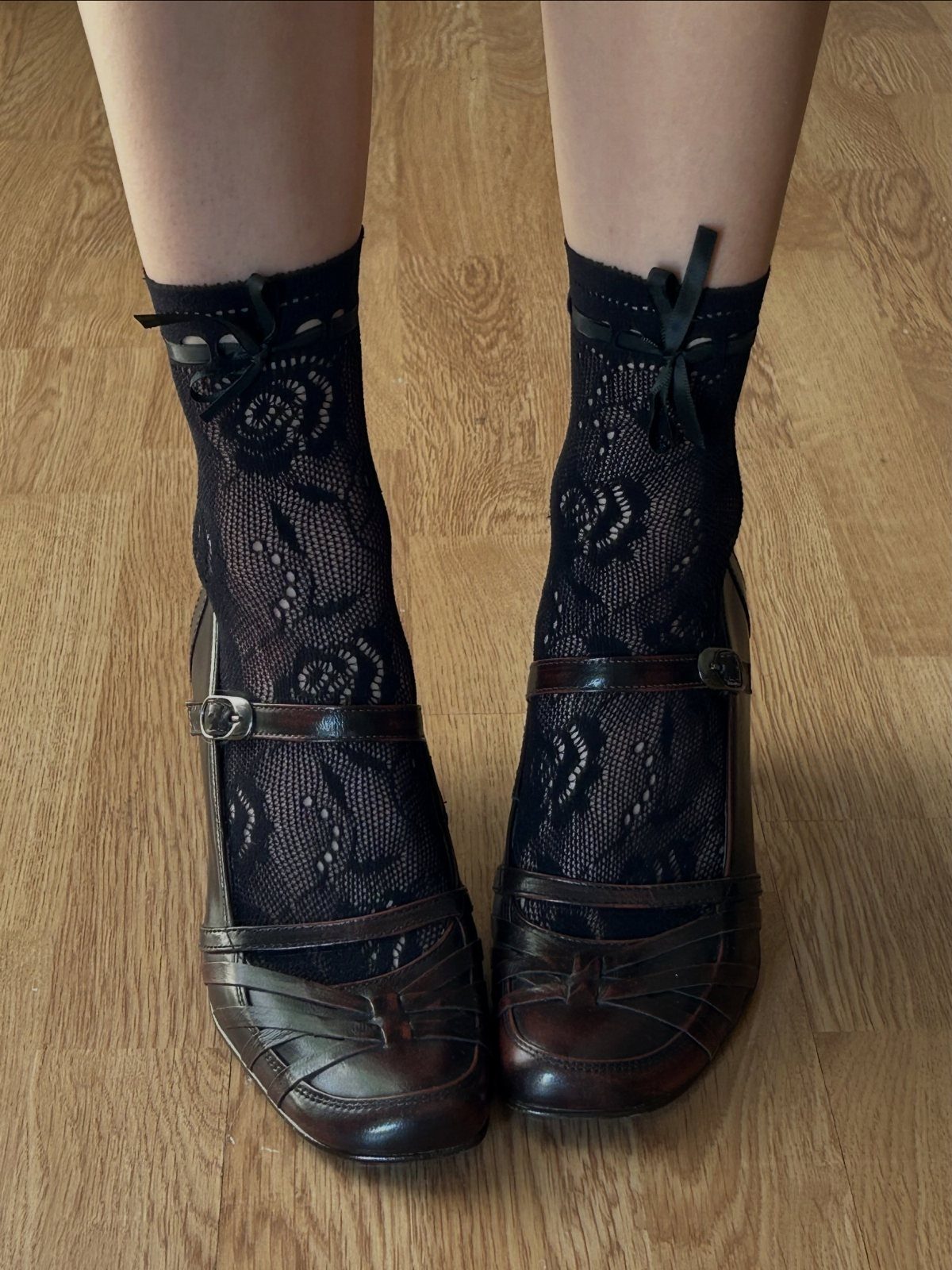 Feet wearing dark brown patent leather Mary Jane heels and black floral lace socks on wood flooring.