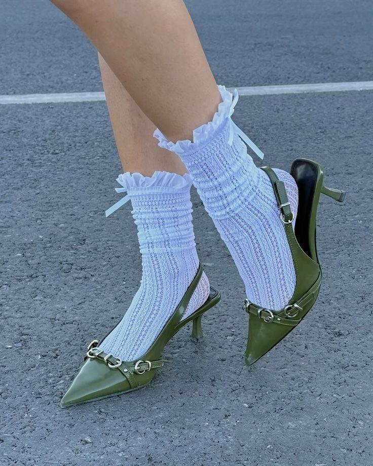 Feet wearing white ruffled lace socks and olive green kitten heels on asphalt.