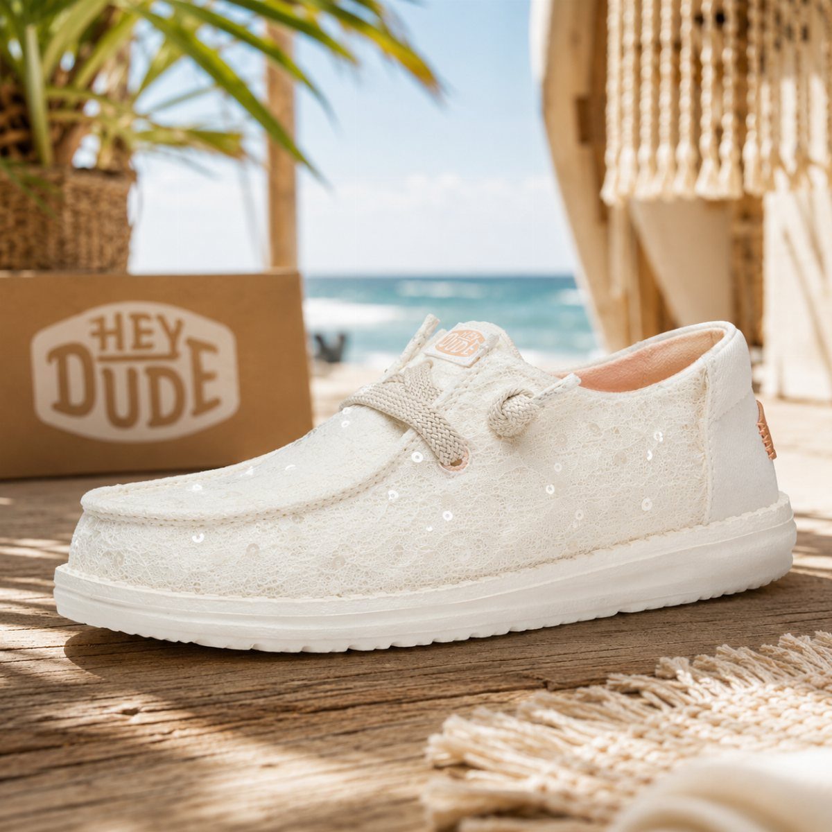 A white sparkly Hey Dude slip-on shoe resting on a wooden deck near a beach.