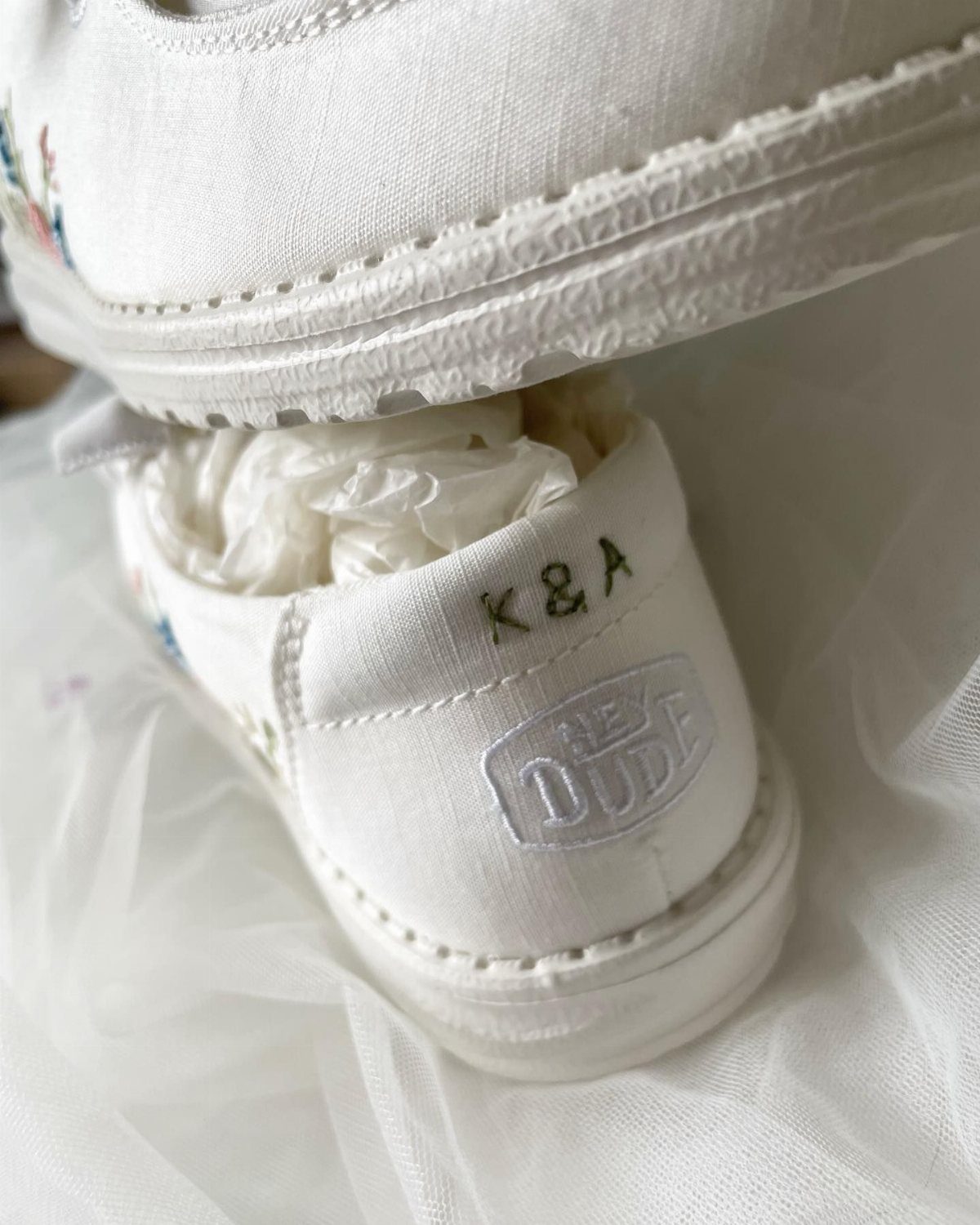 White canvas Hey Dude shoes with embroidered initials K&A on the heel resting on white fabric.