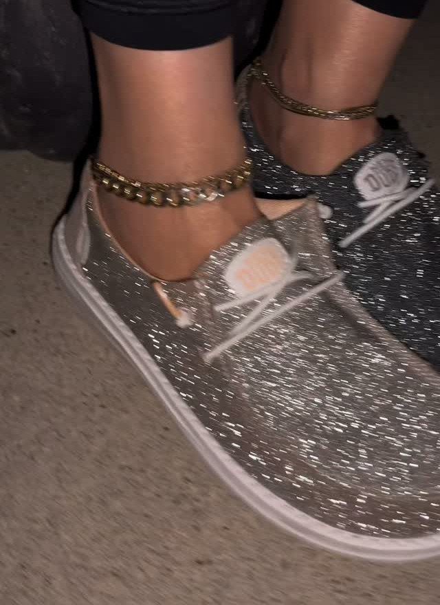 Silver glittery Hey Dude slip-on shoes worn with gold chain anklets on a person's feet.