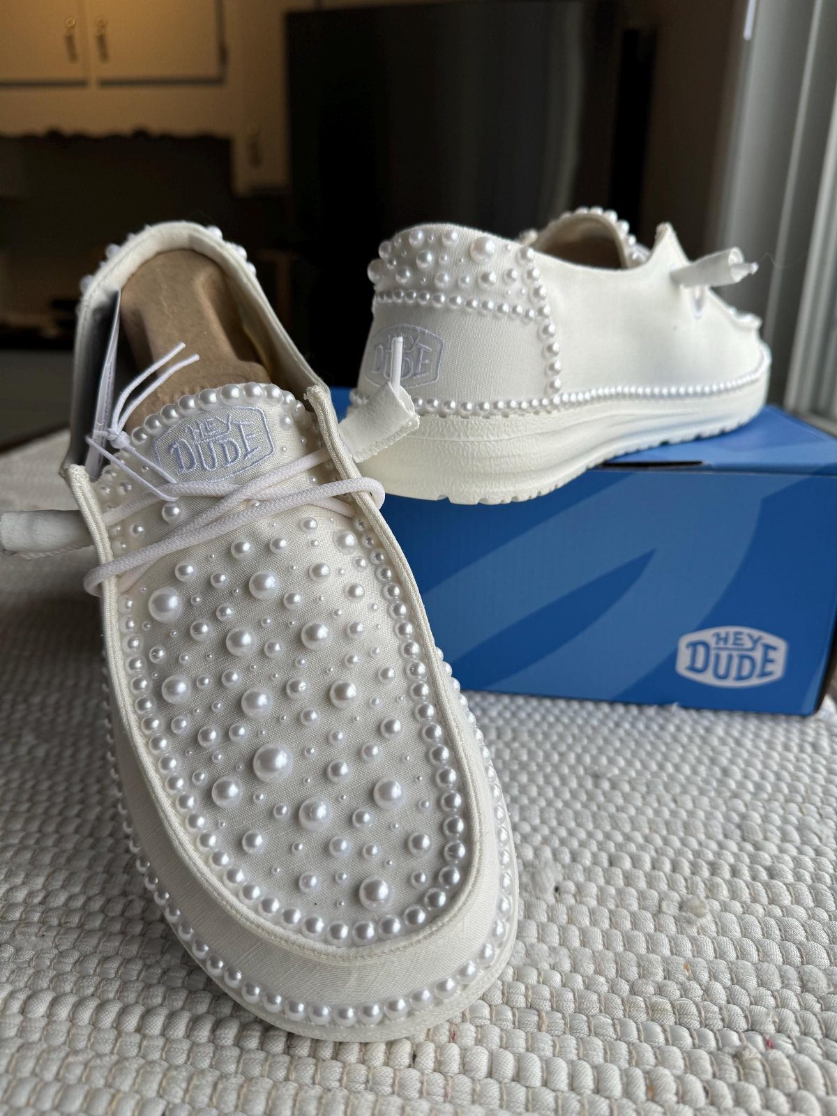 White Hey Dude loafers embellished with white pearls resting next to a blue branded shoebox.