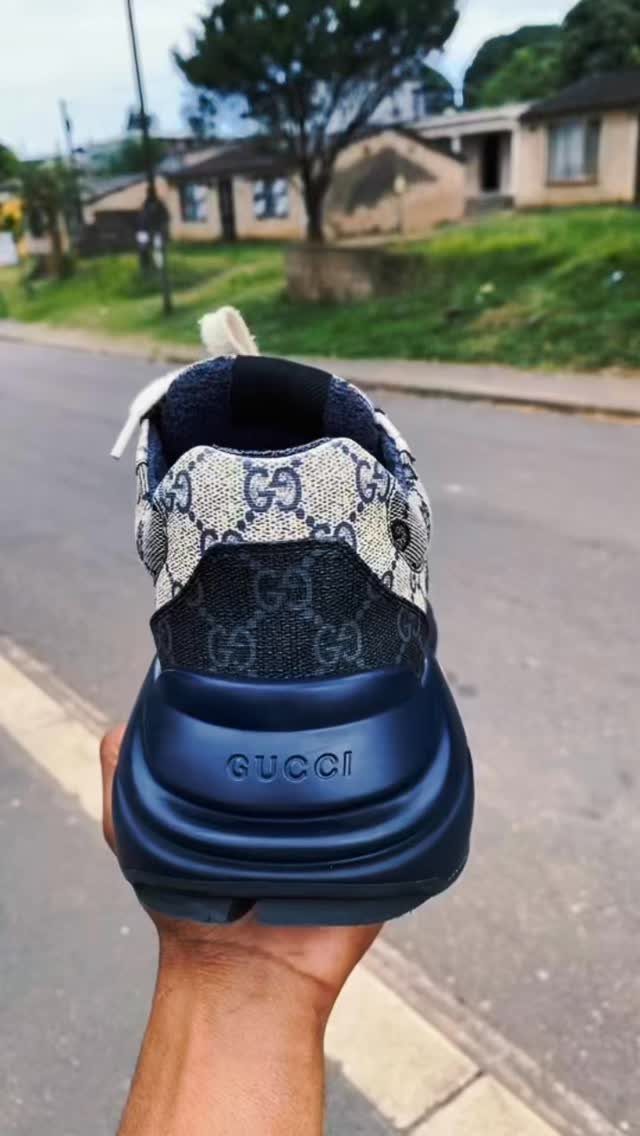 A hand holding a navy and beige Gucci designer sneaker against an outdoor street background.