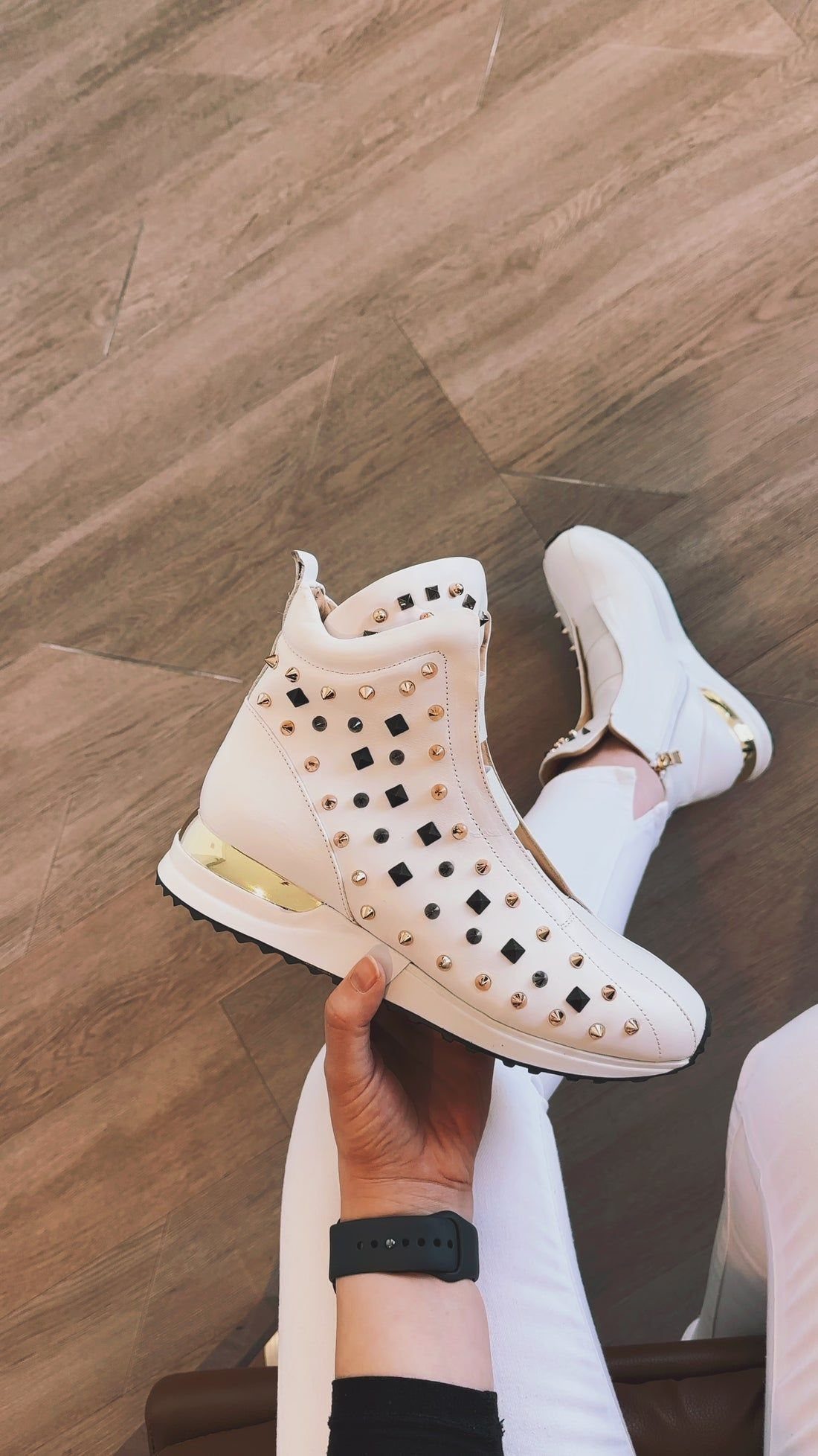 A hand holding a white high-top leather sneaker with gold and black spikes.