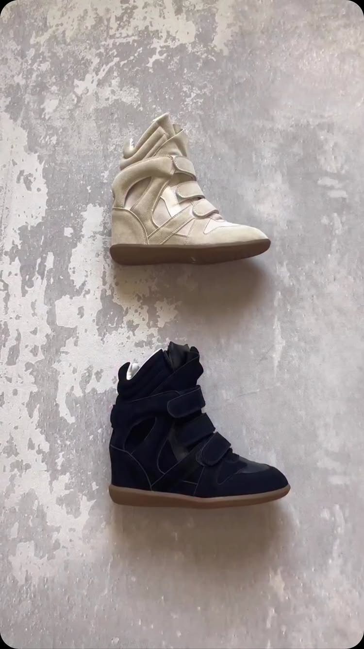 Two high-top velcro sneakers, one cream and one navy, on a grey concrete surface.