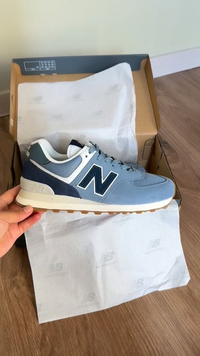 A hand holds a light blue and navy New Balance 574 sneaker over an open shoe box.