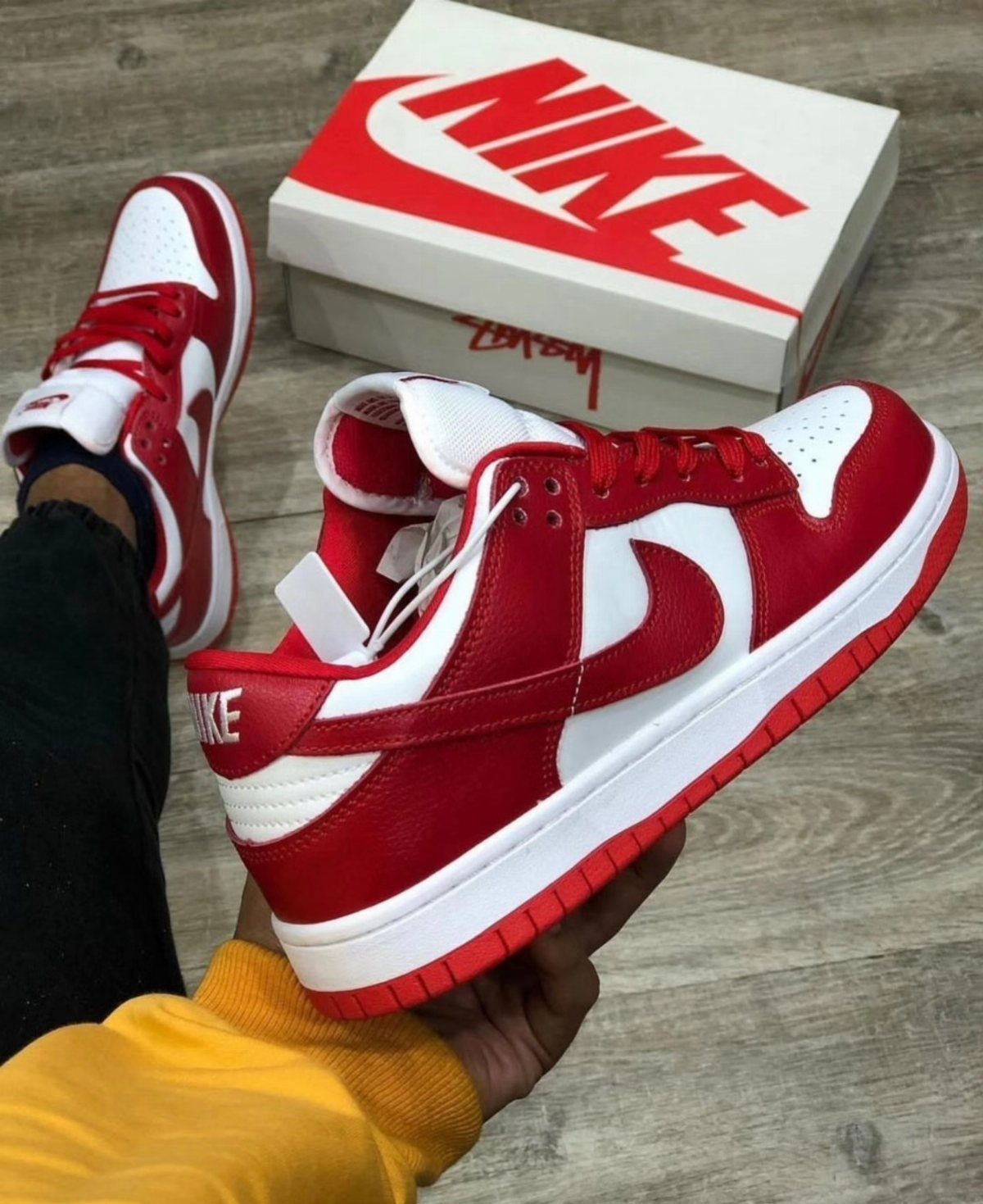 Red and white Nike Dunk Low sneakers held by a hand near a person's foot.
