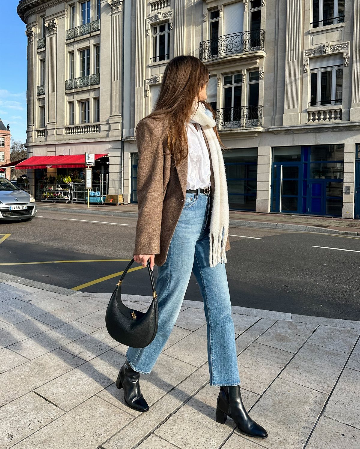 Woman walking in a city wearing a brown blazer, blue jeans, and black ankle boots.