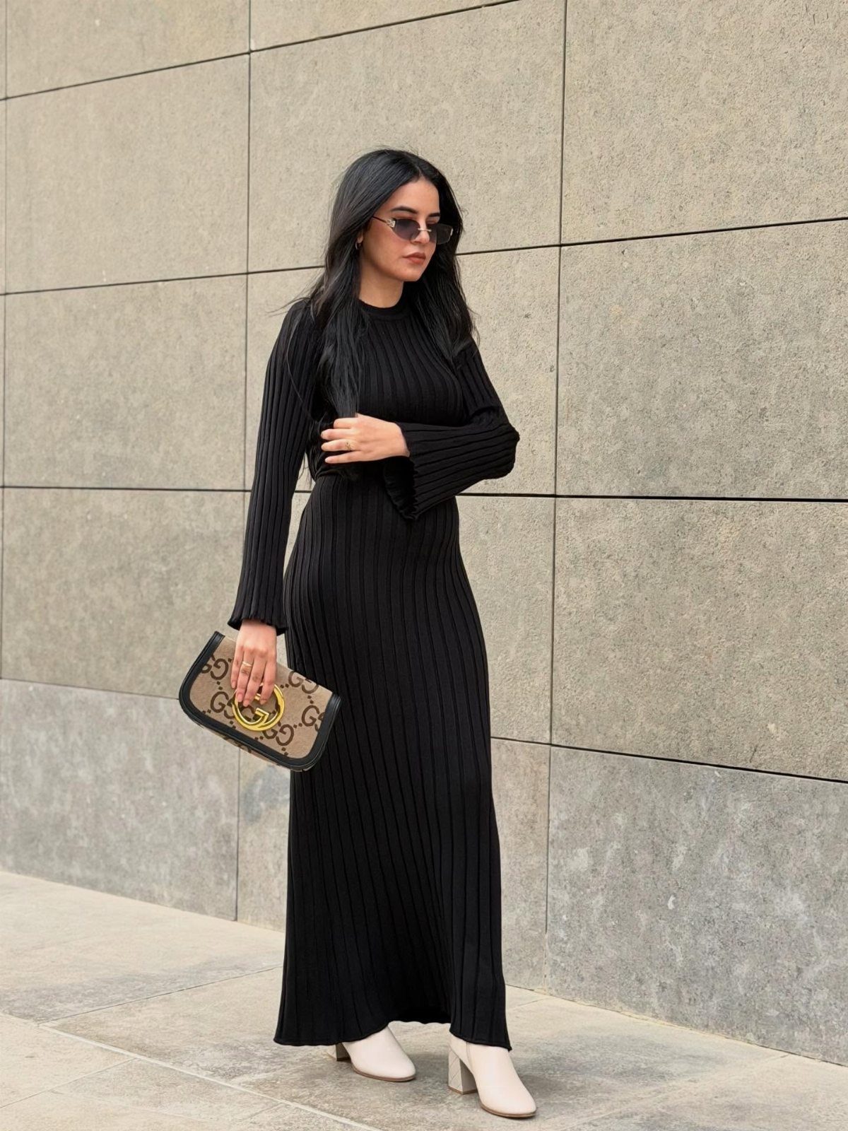 Woman in a black ribbed maxi dress and cream ankle boots standing against a stone wall.