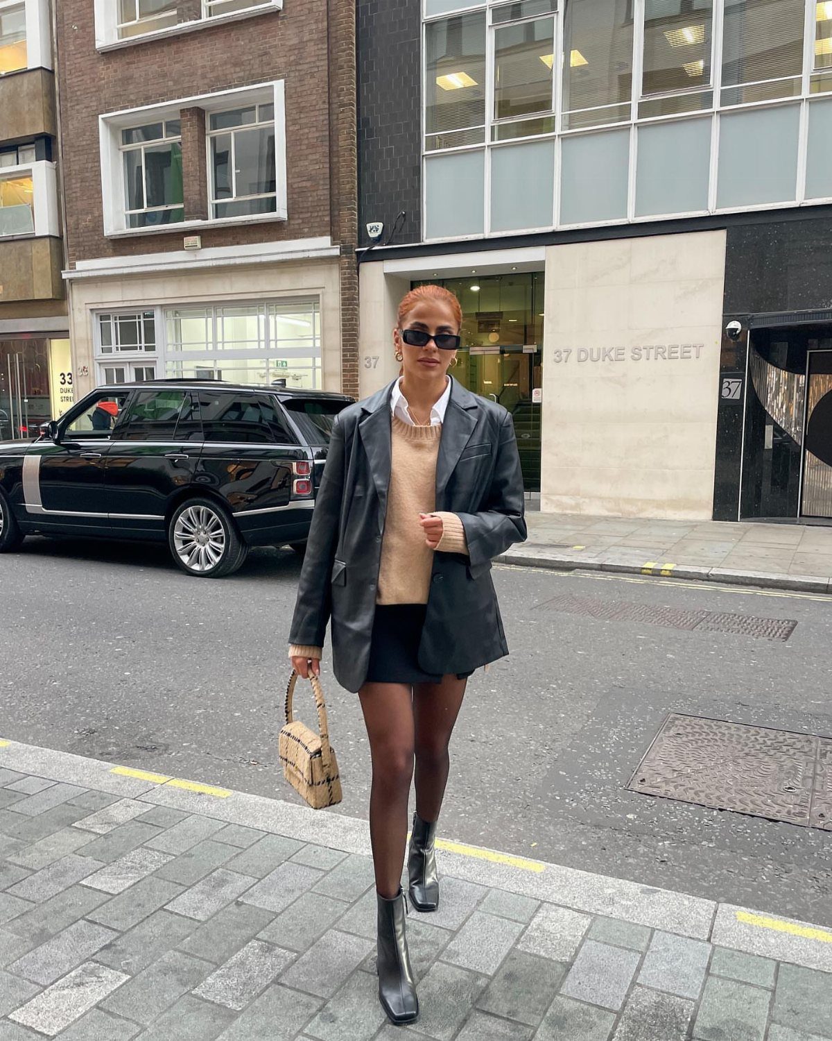A woman in a black blazer and mini skirt posing on a city street wearing black ankle boots.