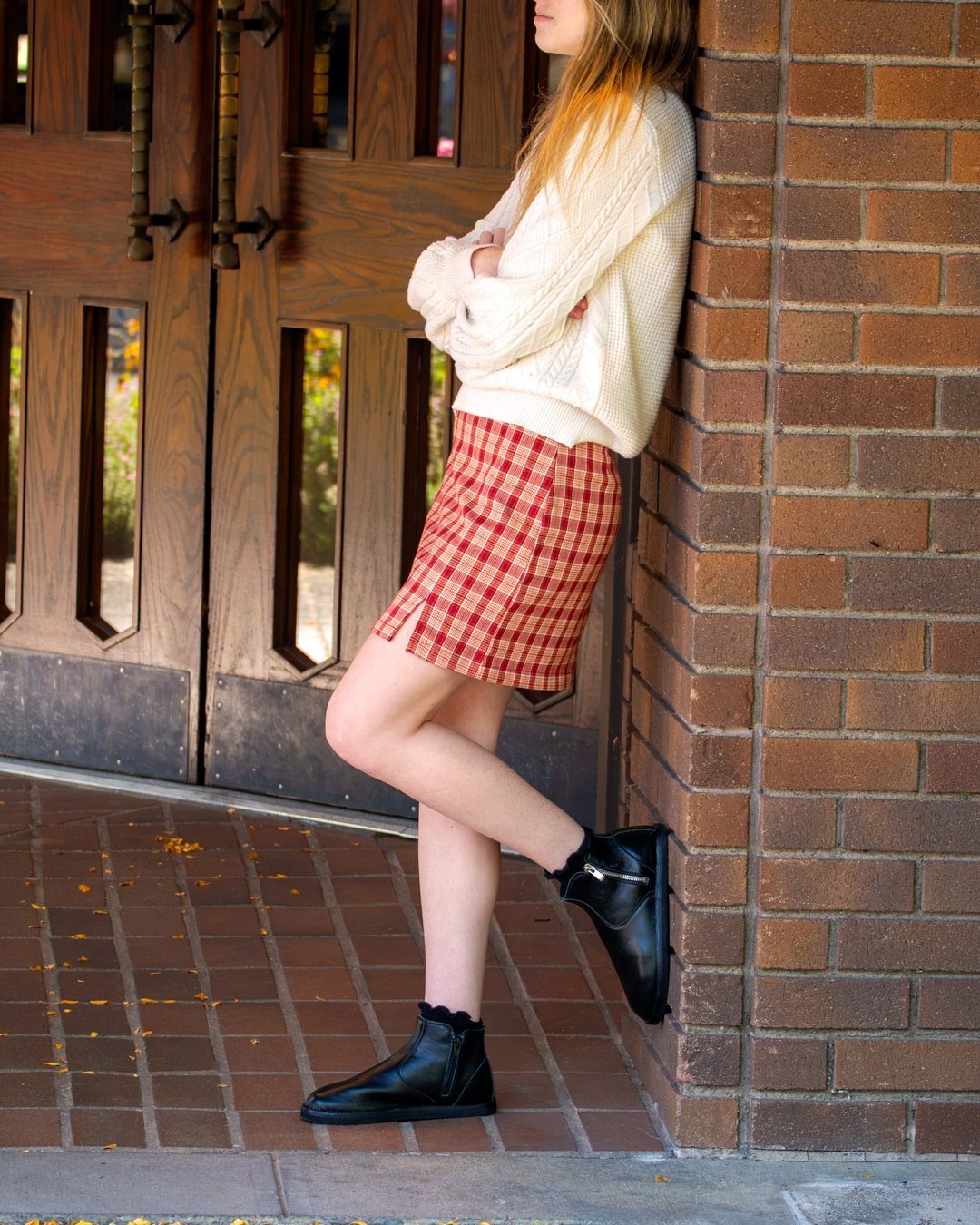 A person wearing a white sweater, red plaid skirt, and black ankle boots leaning against a brick wall.