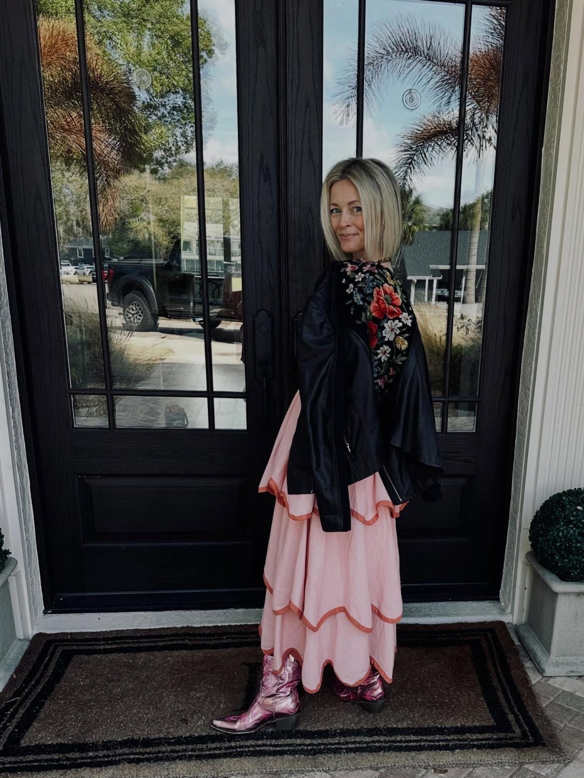 A woman in a pink skirt and metallic pink ankle boots standing before dark doors.