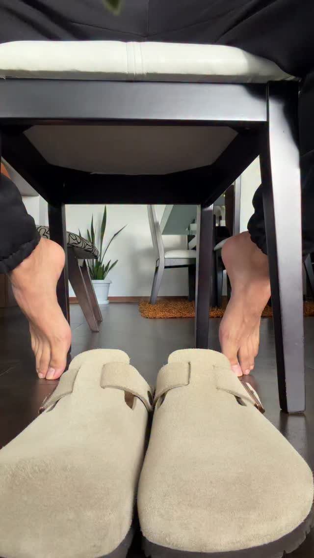 Beige suede Birkenstock clogs on a dark floor with bare feet visible under a chair.