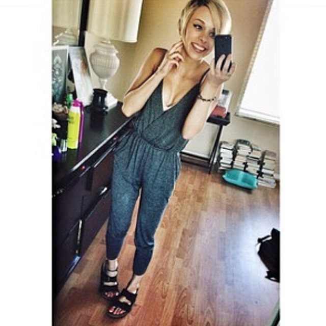 A blonde woman in a grey jumpsuit posing for a mirror selfie wearing black sandals.