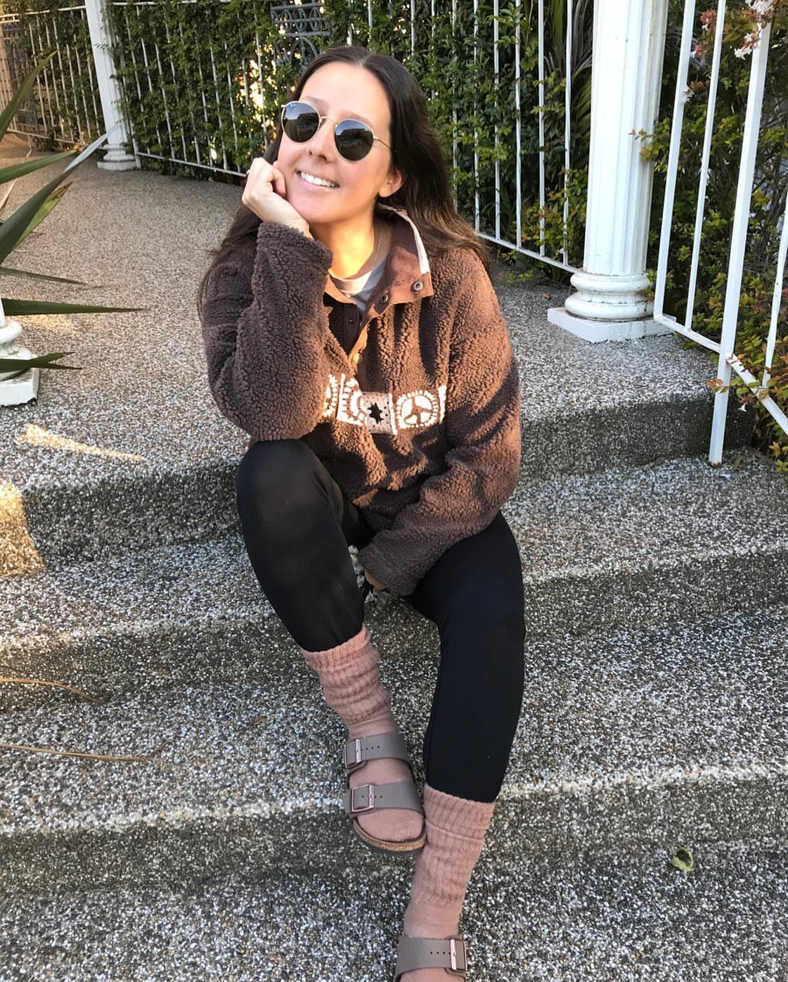 A woman sitting on steps wearing a brown fleece sweatshirt, black leggings, and Birkenstock sandals.