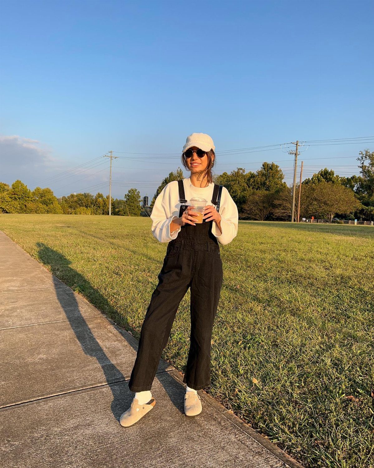 Woman in black overalls and white shirt wearing beige sandals on a sidewalk by a field.