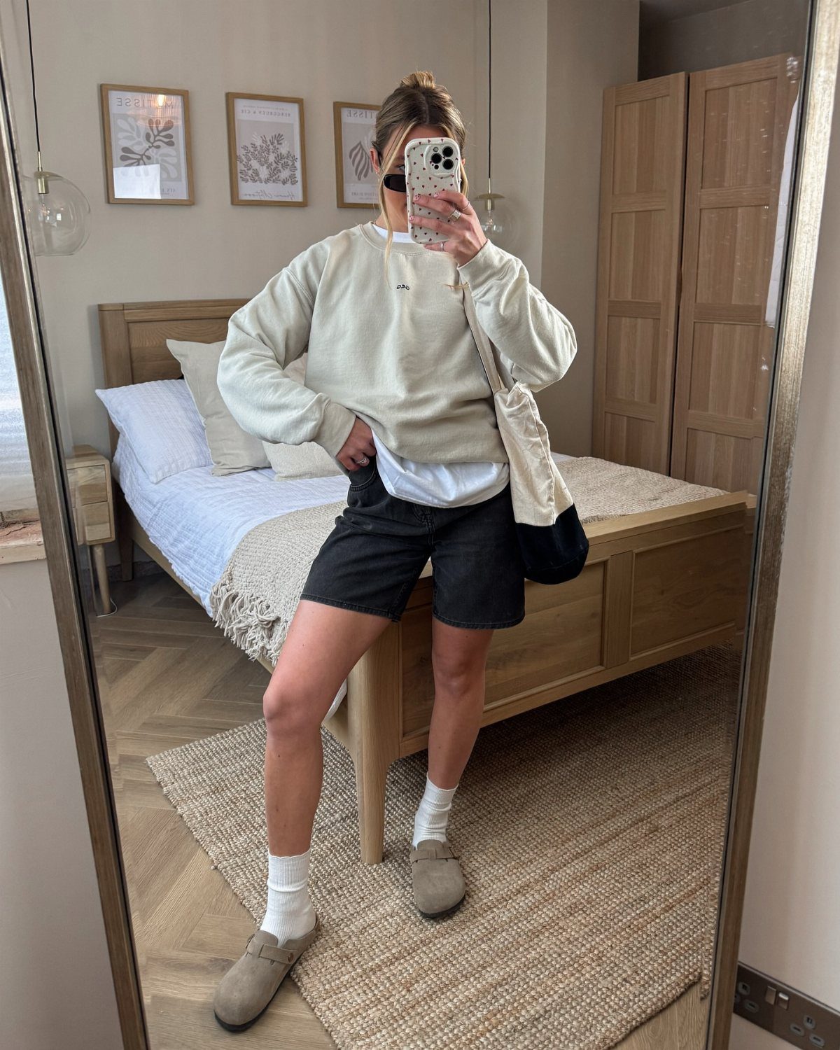 Person wearing cream sweatshirt, black shorts, white socks, and beige clog sandals standing in a bedroom.