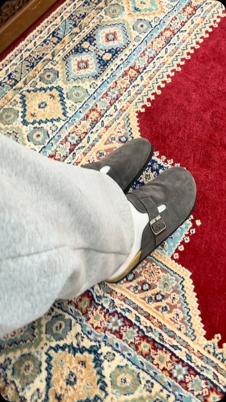Person wearing grey suede sandals with white socks and grey sweatpants on a patterned rug.